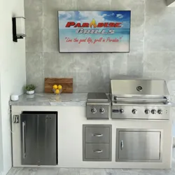 Stainless outdoor kitchen island with grill, mini fridge, and drawers beneath a Paradise Grills TV display