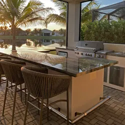 Outdoor kitchen island and bar stools on a covered patio overlooking water and palm trees at sunset