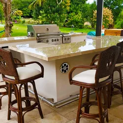 White stucco outdoor kitchen island with granite countertop, grill, and wicker bar stools in tropical backyard