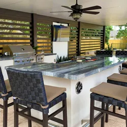 White outdoor kitchen island with blue tile countertop, grill, and woven bar stools under a pergola