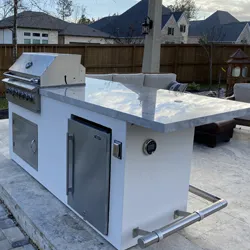 White outdoor kitchen island with grill and refrigerator, set on stone pavers in a residential backyard